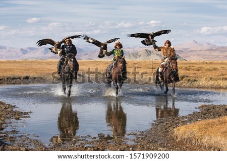 A group of traditional kazakh eagle hunters galloping through the water with their golden eagles. Ulgii, Mongolia.