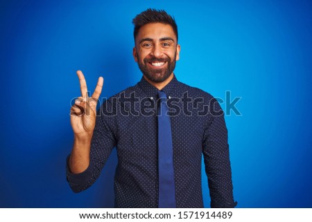Young indian businessman wearing elegant shirt and tie standing over isolated blue background showing and pointing up with fingers number two while smiling confident and happy.