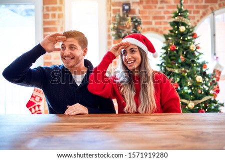 Young couple wearing santa claus hat sitting on chair and table around christmas tree at home very happy and smiling looking far away with hand over head. Searching concept.