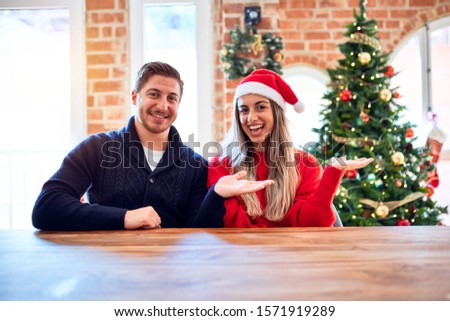 Young couple wearing santa claus hat sitting on chair and table around christmas tree at home smiling cheerful presenting and pointing with palm of hand looking at the camera.