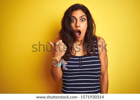 Young beautiful woman wearing striped t-shirt standing over isolated yellow background Surprised pointing with hand finger to the side, open mouth amazed expression.