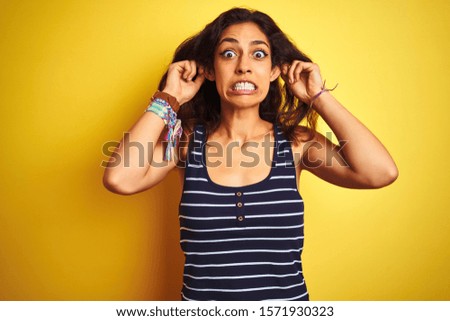 Young beautiful woman wearing striped t-shirt standing over isolated yellow background Smiling pulling ears with fingers, funny gesture. Audition problem
