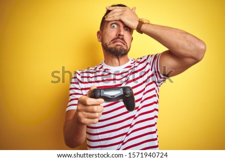 Young handsome man playing video games using joystick game pad over yellow background stressed with hand on head, shocked with shame and surprise face, angry and frustrated. Fear and upset for mistake