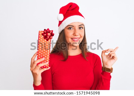 Young beautiful girl wearing Christmas Santa hat holding gift over isolated white background very happy pointing with hand and finger to the side