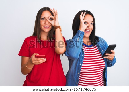 Young beautiful women using smartphone standing over isolated white background with happy face smiling doing ok sign with hand on eye looking through fingers
