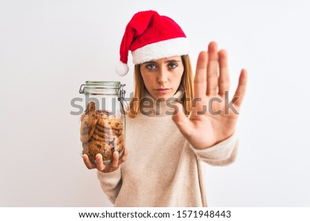Beautiful redhead woman wearing christmas hat holding cookies jar over isolated background with open hand doing stop sign with serious and confident expression, defense gesture