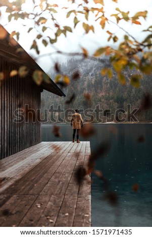 Full body shot of young male photographer in brown coat taking pictures with a small mirrorless camera standing on a lake house deck in lake plansee, Austria / During Autumn with falling leaves 