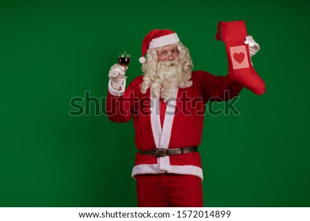Emotional male actor in a costume of Santa Claus holds a glass of wine and a Christmas sock for gifts in his hands and poses on a green chrome background
