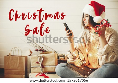 Christmas Sale text sign on stylish happy girl in santa hat looking at phone screen in festive christmas lights on background of  presents. Special discount offer. Holiday shopping online