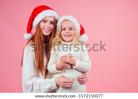 redhaired ginger woman with small little blond girl wearing santas hat holding many gift boxes with whire rat in studio pink background