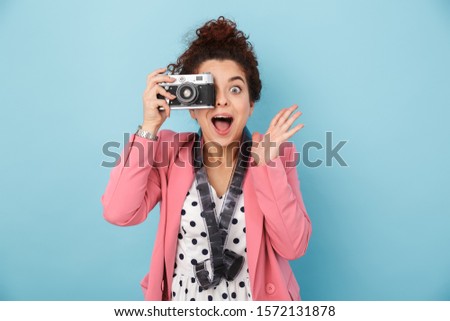 Cheerful excited young girl standing isolated over blue background, taking picture with photo camera