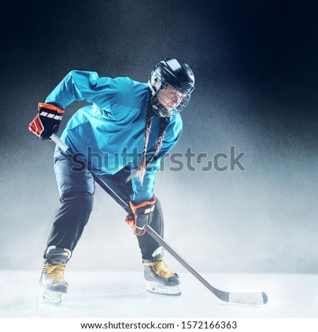 Young female hockey player with the stick on ice court and blue background. Sportswoman wearing equipment and helmet practicing. Concept of sport, healthy lifestyle, motion, movement, action.