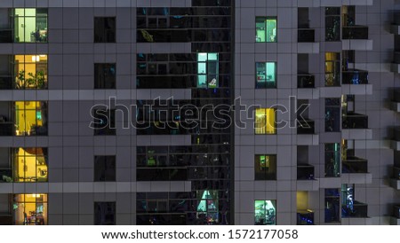 Rows of glowing windows with people in the interior of apartment building at night. Modern skyscraper from glass and concrete. Concept for business and modern life. Zoom out