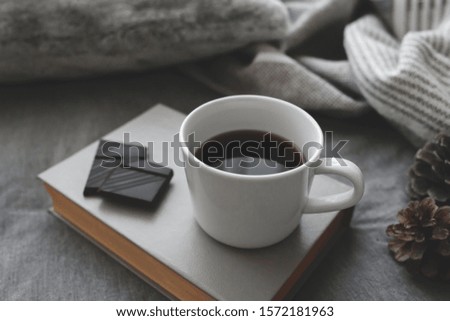 Cup of coffee, chocolate, book, blanket, plaid and pine cones lie on the bed with gray linens. Still life details. Winter scene. Cozy concept. Dark grey colors. Close up, selective focus.