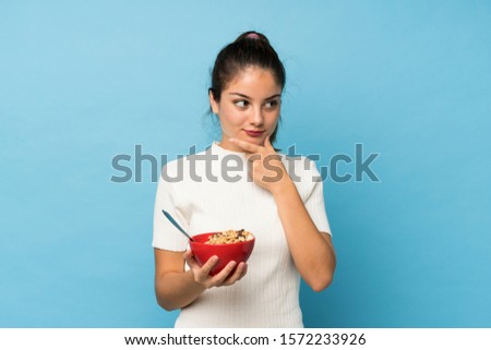 Young brunette girl over isolated blue background holding a bowl of cereals and thinking