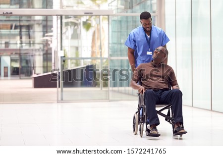 Male Nurse Wearing Scrubs Wheeling Patient In Wheelchair Through Lobby Of Modern Hospital Building Royalty-Free Stock Photo #1572241765