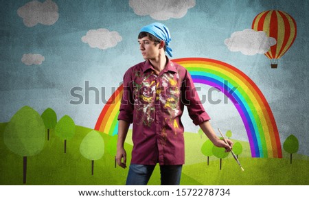 Young male artist with paintbrush. Happy painter in shirt and bandana standing on background colorful picture on wall. Summer landscape with hot air balloon and rainbow. Creative hobby and profession