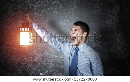 Screaming businessman holding glowing lantern on background grunge wall. Front view of young emotional man in shirt and tie looking for something in dark. Scared business person walking with lantern