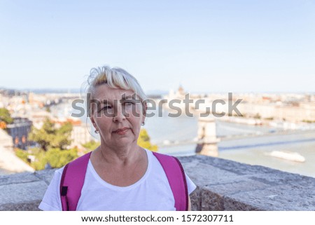 Adult blonde woman in a white T-shirt, with a crimson color backpack look at the camera against the background of the old city of Budapest. Travel vacation city concept