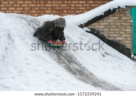 Funny child dressed in adult oversized jacket riding sled down icey trek from cellar in winter day. Little girl with emotional face riding slide in the village yard