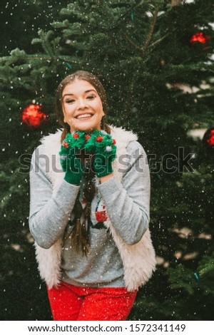 Festive portrait of happy young woman posing outside by the Christmas tree, wearing green mittens