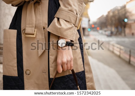 street style fashion details. close up, young fashion blogger wearing autumn trench coat and a white and golden black analog wrist watch. checking the time, holding a beautiful brown leather purse.
 Royalty-Free Stock Photo #1572342025