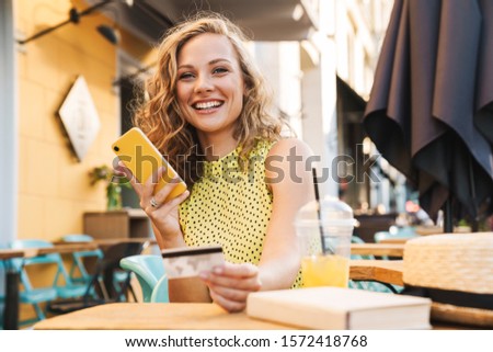 Photo of a young amazing smiling woman outdoors in cafe using mobile phone.