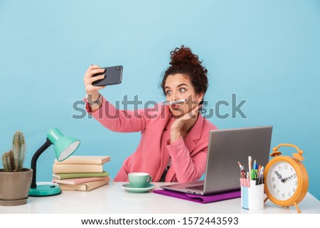 Image of amusing woman taking selfie photo on cellphone and grimacing while working at desk isolated over blue background