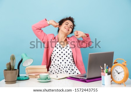 Image of sleepy woman stretching her body with eyes closed while working at desk isolated over blue background