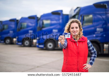 Woman truck driver proudly holding commercial driving license. In background parked trucks. Truck driving school and job openings for new drivers. Royalty-Free Stock Photo #1572451687