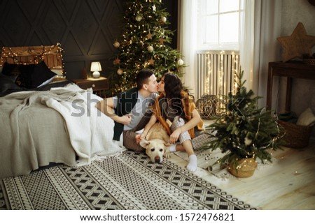 happy couple is sitting near the bed with a dog, christmas. new year at home. family and date. hearth, home clothes and sweaters. comfort and warmth of hearts