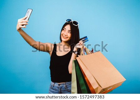 Portrait of an excited beautiful girl wearing dress and sunglasses holding shopping bags.Happy Asian woman taking selfie photo with smartphone while holding shopping bags. black friday season sale