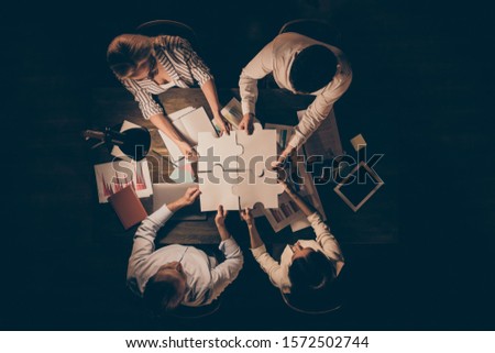 High angle above view vertical photo of four business people colleagues sitting table circle working late night holding paper puzzle pieces came to unity formalwear indoors