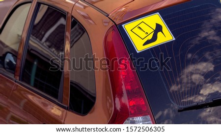 The main object is a sign on the glass of a car, a yellow square with women's shoes. Sign - attention, female driver. Blurred background. Passenger car in red.