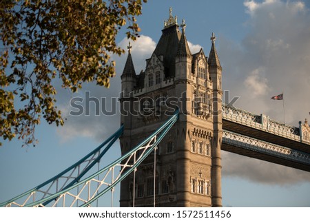 View of a section of the Tower Bridge under a blue sky before sunset in London, England, United Kingdom, with trees in the foreground