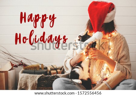 Happy Holidays text sign on cute cat playing with santa hat on stylish happy girl in festive christmas lights on background of modern presents. Seasons greeting card