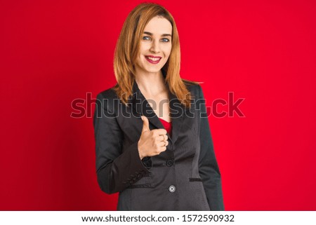 Young caucasian business woman wearing a suit over isolated red background doing happy thumbs up gesture with hand. Approving expression looking at the camera with showing success.