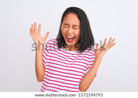 Young chinese woman wearing striped t-shirt standing over isolated white background celebrating mad and crazy for success with arms raised and closed eyes screaming excited. Winner concept