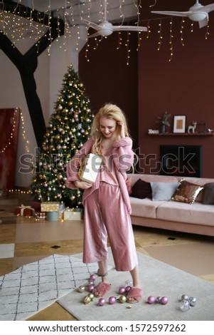 The concept of a Christmas party. Beautiful cheerful happy blonde with dreadlocks in pink home pajamas holds and plays with a gift in her hand on the background of the Christmas tree and the fireplace