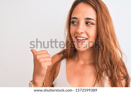 Young redhead woman stading over white isolated background pointing and showing with thumb up to the side with happy face smiling