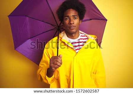 Afro american man wearing rain coat and umbrella standing over isolated yellow background with a confident expression on smart face thinking serious