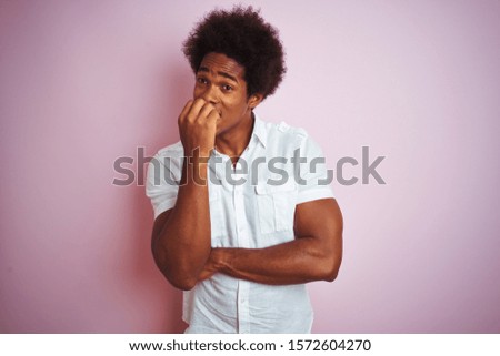Young american man with afro hair wearing white shirt standing over isolated pink background looking stressed and nervous with hands on mouth biting nails. Anxiety problem.
