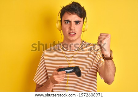 Teenager boy playing video games using gamepad over isolated yellow background annoyed and frustrated shouting with anger, crazy and yelling with raised hand, anger concept