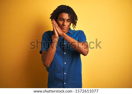 Afro man with dreadlocks wearing casual denim shirt standing over isolated yellow background sleeping tired dreaming and posing with hands together while smiling with closed eyes.