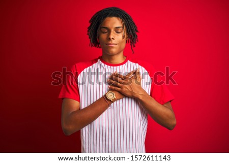 Afro man with dreadlocks wearing striped t-shirt standing over isolated red background smiling with hands on chest with closed eyes and grateful gesture on face. Health concept.