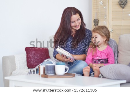 Mom and daughter are sitting on the sofa at home and reading a book. Happy loving family. Mother and her daughter child girl playing. Good time at home.