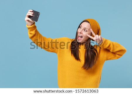Funny young brunette woman girl in sweater hat posing isolated on blue background studio portrait. People lifestyle concept. Mock up copy space. Doing selfie shot on mobile phone shwoing victory sign