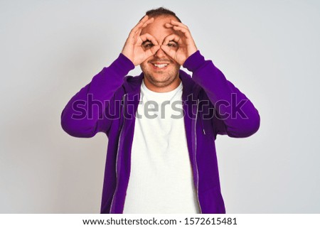 Young man wearing purple sweatshirt standing over isolated white background doing ok gesture like binoculars sticking tongue out, eyes looking through fingers. Crazy expression.