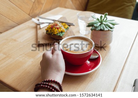 Hipster urban style Coffee shop with fresh green breakfast on wooden table. Young woman hand reaching for cappuccino coffee and heathy avocado toast styled with plants. Bright spacious flat lay style.