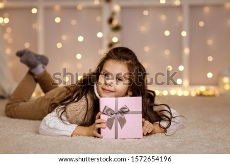 Happy little child girl with gift box sitting on the floor on the background with lights. Merry Christmas. copy space